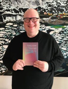 Jonathan standing in front of a painting, smiling, holding a copy of Diminished Faculties