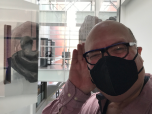 Jonathan (a balding middle aged white man in chunky glasses) in a black KN-95 mask, cupping his ear and leaning in the direction of a window reflection of a sculpture of an ear.